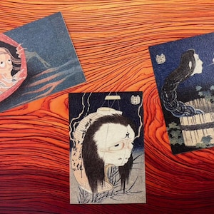 以下が含まれることがあります： 伝統的な幽霊のイメージを特徴とする3つの日本の版画。暗く、雰囲気のあるスタイルで、さまざまな幽霊の姿を描いています。鮮やかな赤みがかったオレンジ色の木目調の背景に配置されたアートワーク。