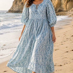 May include: A light blue paisley print dress with a round neckline and three-quarter sleeves. The dress has a flowing skirt and is worn on a sandy beach. The dress is a midi length.