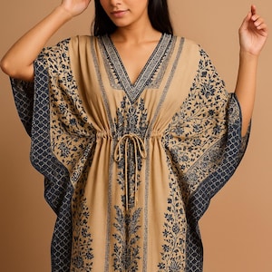 A tan caftan with a V-neck and a drawstring waist. The caftan features a dark black floral pattern and trim along the neckline, sleeves, and hem. The sleeves are wide and flowing. The caftan is made of a lightweight fabric.