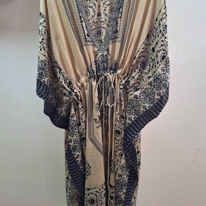 May include: A beige kaftan with a V-neck and a drawstring waist. The garment features a dark black floral and geometric pattern along the neckline, sleeves, and hem. The kaftan is hanging on a wooden hanger.
