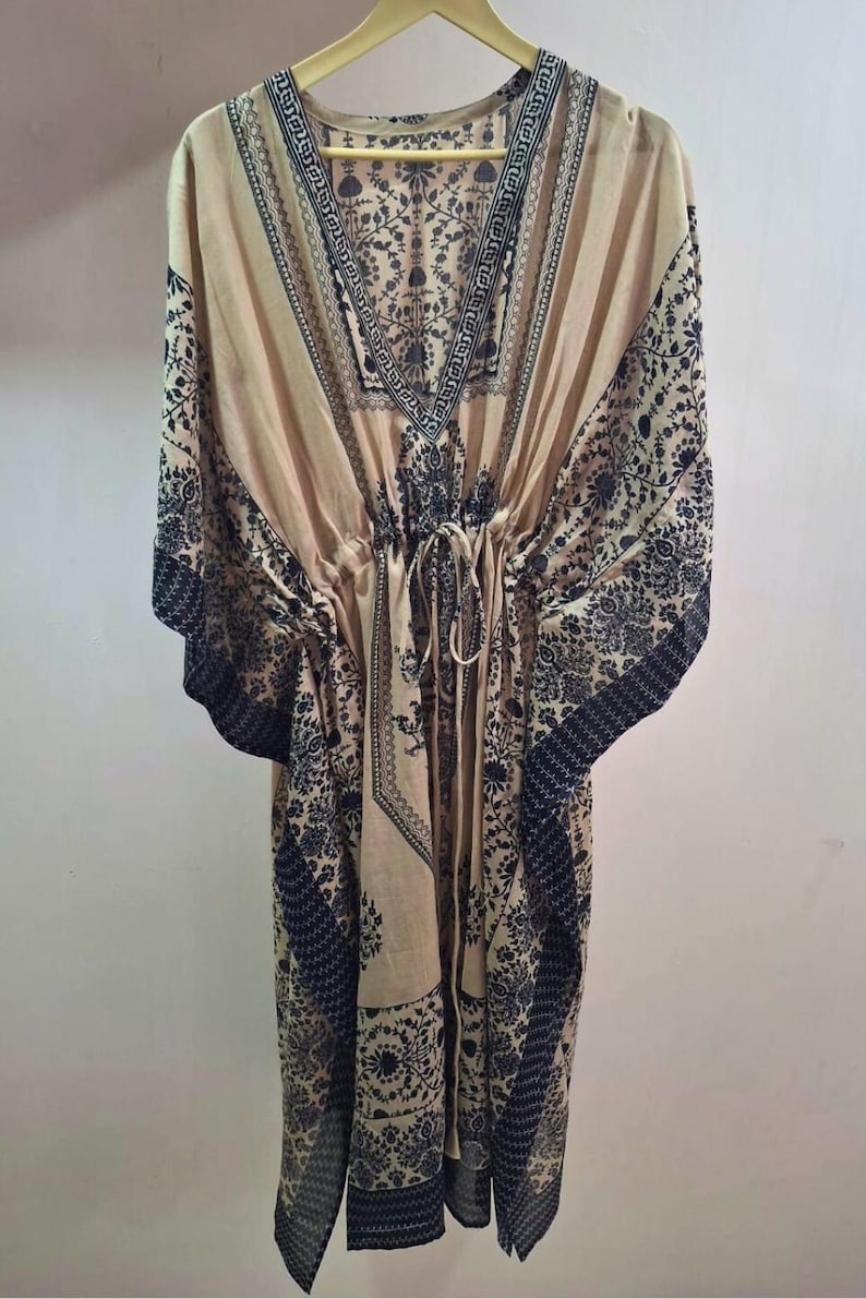 May include: A beige kaftan with a V-neck and a drawstring waist. The garment features a dark black floral and geometric pattern along the neckline, sleeves, and hem. The kaftan is hanging on a wooden hanger.