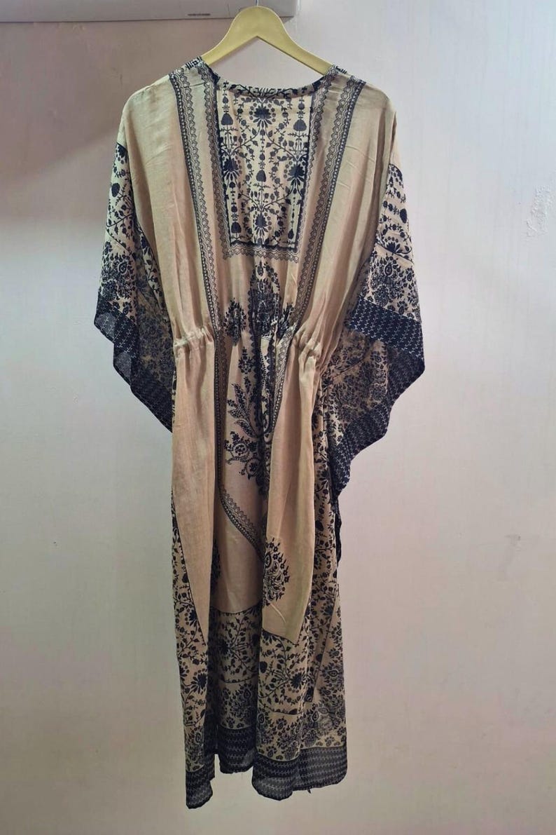 May include: A beige kaftan dress with a black floral pattern. The dress has a drawstring waist and wide sleeves. The fabric appears lightweight and flowing, suitable for warm weather.