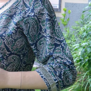 Puede incluir: Vestido azul marino con estampado de cachemira en verde y blanco. El vestido presenta un escote en V y mangas tres cuartos con ribete dorado. La tela tiene un patr&oacute;n detallado y repetitivo.
