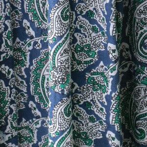 Puede incluir: Primer plano de una tela con un patr&oacute;n de cachemira repetido. El dise&ntilde;o presenta elementos blancos, verdes y azules. La tela est&aacute; drapeada, mostrando los pliegues y la textura del material. La est&eacute;tica general es un textil cl&aacute;sico y detallado.