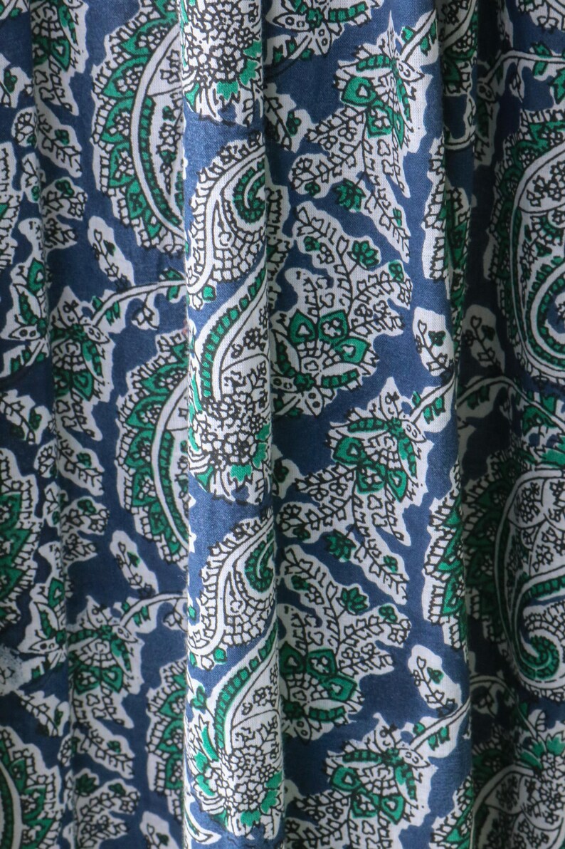 Puede incluir: Primer plano de una tela con un patr&oacute;n de cachemira repetido. El dise&ntilde;o presenta elementos blancos, verdes y azules. La tela est&aacute; drapeada, mostrando los pliegues y la textura del material. La est&eacute;tica general es un textil cl&aacute;sico y detallado.
