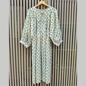 May include: A cream-colored dress with a blue floral pattern. The dress has long, puffy sleeves and a gathered waist. It hangs on a wooden hanger against a slatted background. The dress is knee-length.