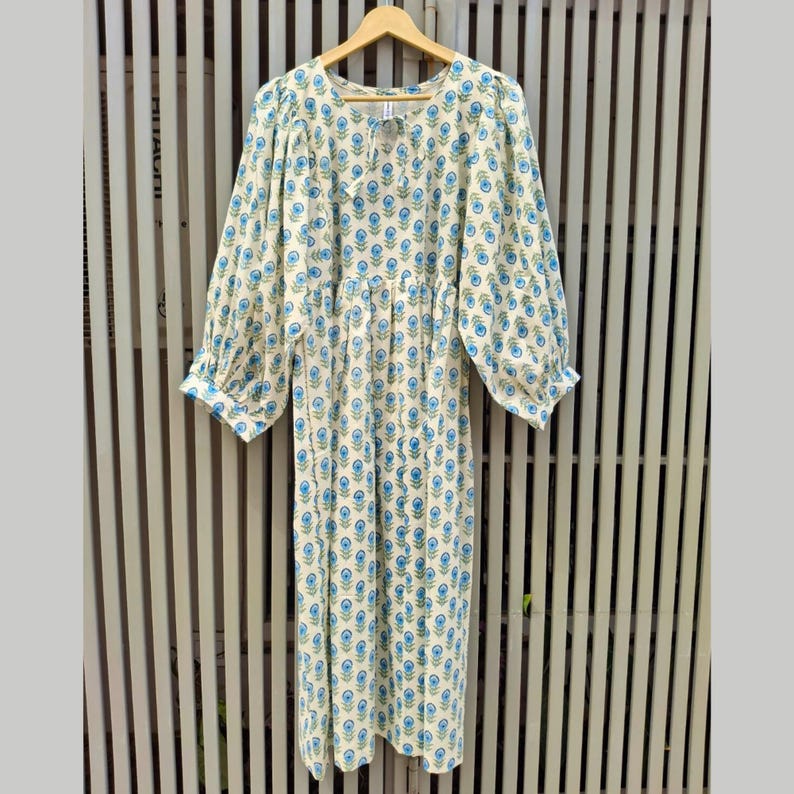 May include: A cream-colored dress with a blue floral pattern. The dress has long, puffy sleeves and a gathered waist. It hangs on a wooden hanger against a slatted background. The dress is knee-length.