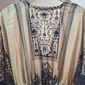 May include: A beige tunic with a dark black floral pattern and decorative trim. The tunic is hanging on a wooden hanger. The fabric appears lightweight and flowing, suitable for warm weather.