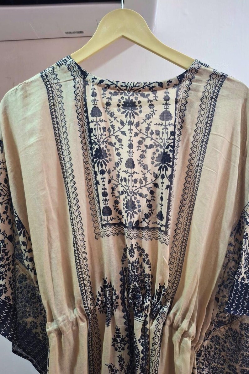May include: A beige tunic with a dark black floral pattern and decorative trim. The tunic is hanging on a wooden hanger. The fabric appears lightweight and flowing, suitable for warm weather.