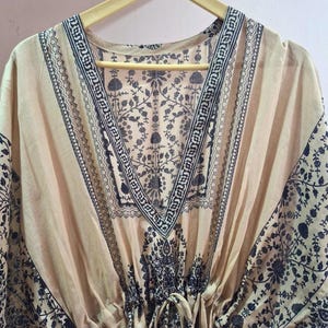 May include: A beige kaftan dress with a V-neck and a drawstring waist. The dress features a dark black floral pattern along the neckline, sleeves, and hem. The fabric appears lightweight and flowing, suitable for warm weather.