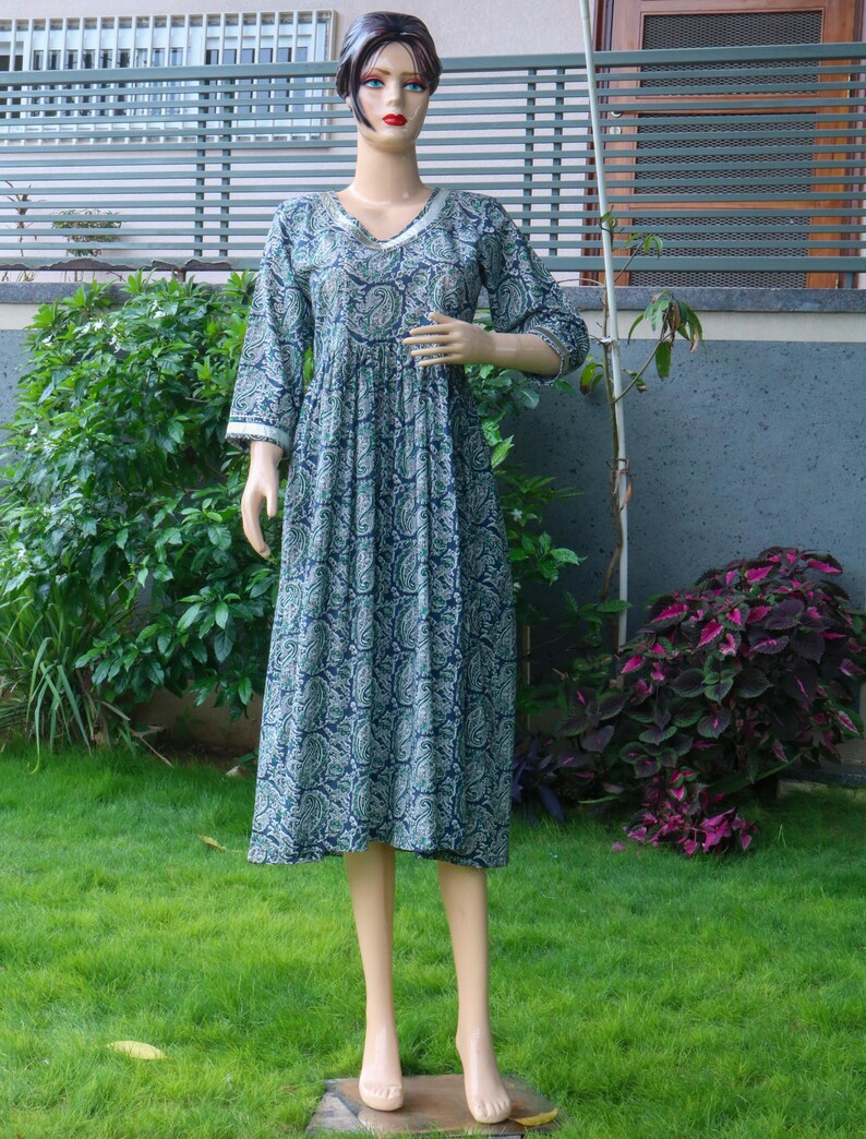 Puede incluir: Vestido largo sobre un maniqu&iacute;. El vestido es de color verde azulado oscuro con un estampado de cachemira en tonos verdes y blancos. Tiene un escote en V y mangas tres cuartos con ribete blanco. El vestido est&aacute; fruncido en la cintura y llega a la pantorrilla.