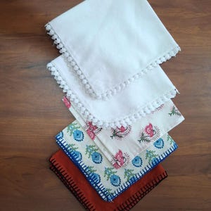 Puede incluir: Una pila de cuatro servilletas cuadradas de tela. Las dos superiores son blancas con ribete de encaje. La tercera servilleta tiene un fondo crema con estampados florales rosas y azules. La servilleta inferior es naranja con costuras negras.
