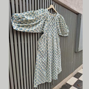 May include: A cream-colored dress with a blue floral pattern and puffy sleeves hangs on a wooden hanger. The dress has a round neckline and a gathered waist. The fabric is lightweight and appears to be cotton or a similar material.