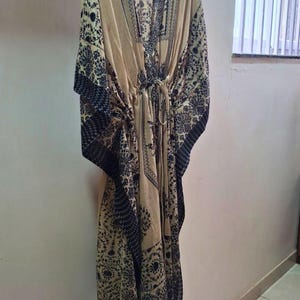 May include: A beige kaftan with a V-neck and a drawstring waist. The kaftan features a dark black floral pattern along the edges and sleeves. The garment is hanging on a wooden hanger against a white wall.