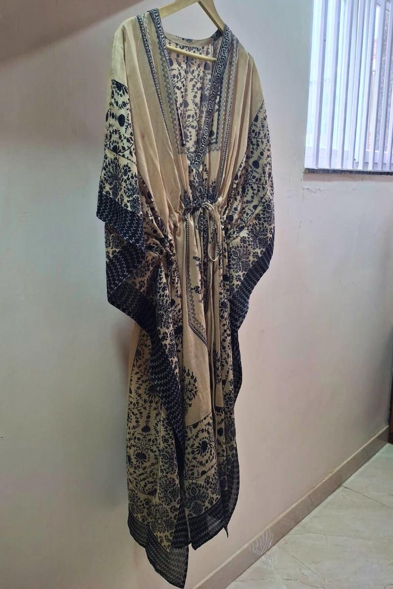 May include: A beige kaftan with a V-neck and a drawstring waist. The kaftan features a dark black floral pattern along the edges and sleeves. The garment is hanging on a wooden hanger against a white wall.