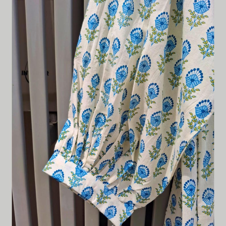 May include: Close-up of a white fabric with a repeating blue floral pattern and green leaves. The fabric is gathered at the cuff of a long sleeve. The floral design is a stylized, symmetrical flower.