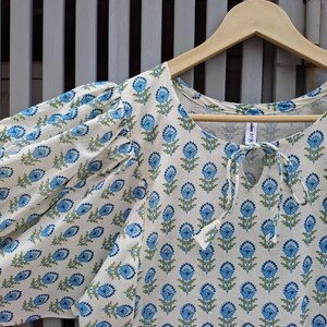 May include: A white blouse with a blue floral pattern and short, puffed sleeves. The blouse has a keyhole neckline with a tie closure and is hanging on a wooden hanger.