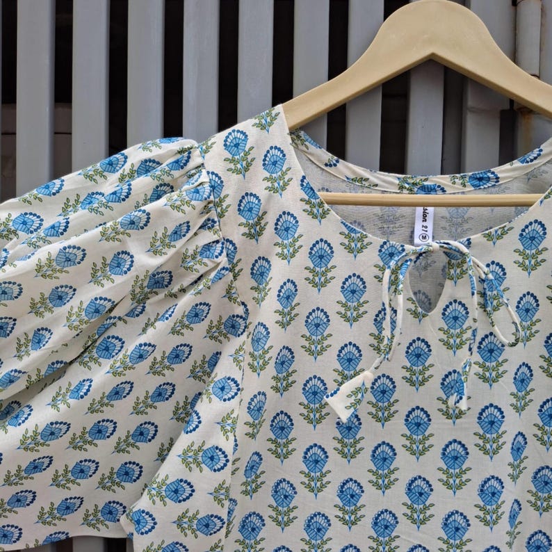 May include: A white blouse with a blue floral pattern and short, puffed sleeves. The blouse has a keyhole neckline with a tie closure and is hanging on a wooden hanger.