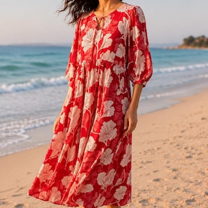 May include: A red midi dress with a white floral pattern. The dress has long sleeves and a tie-neck detail. The model is standing on a sandy beach with the ocean in the background. The dress is a flowy, casual style.