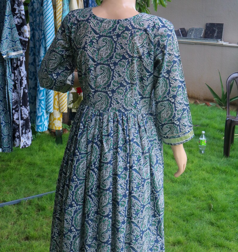 Puede incluir: Un vestido largo azul marino con un estampado de cachemira en tonos verdes y blancos. El vestido tiene mangas tres cuartos y cintura fruncida. El vestido se exhibe en un maniqu&iacute; al aire libre.