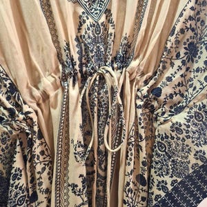 May include: A beige kaftan with a V-neck and a drawstring waist. The fabric is adorned with a dark black floral and paisley pattern. The sleeves and neckline are trimmed with a decorative border.