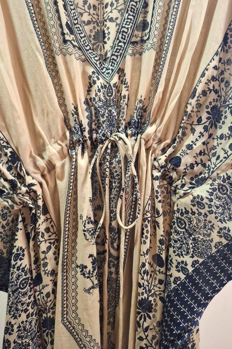 May include: A beige kaftan with a V-neck and a drawstring waist. The fabric is adorned with a dark black floral and paisley pattern. The sleeves and neckline are trimmed with a decorative border.