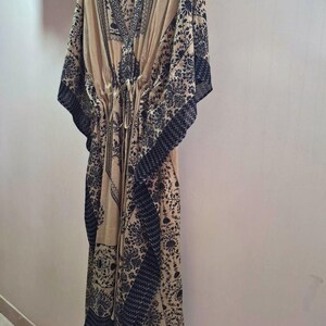 May include: A beige kaftan dress with a V-neck and a drawstring waist. The dress features a dark black floral pattern along the edges, sleeves, and neckline. It is hanging on a wooden hanger against a light-colored wall.
