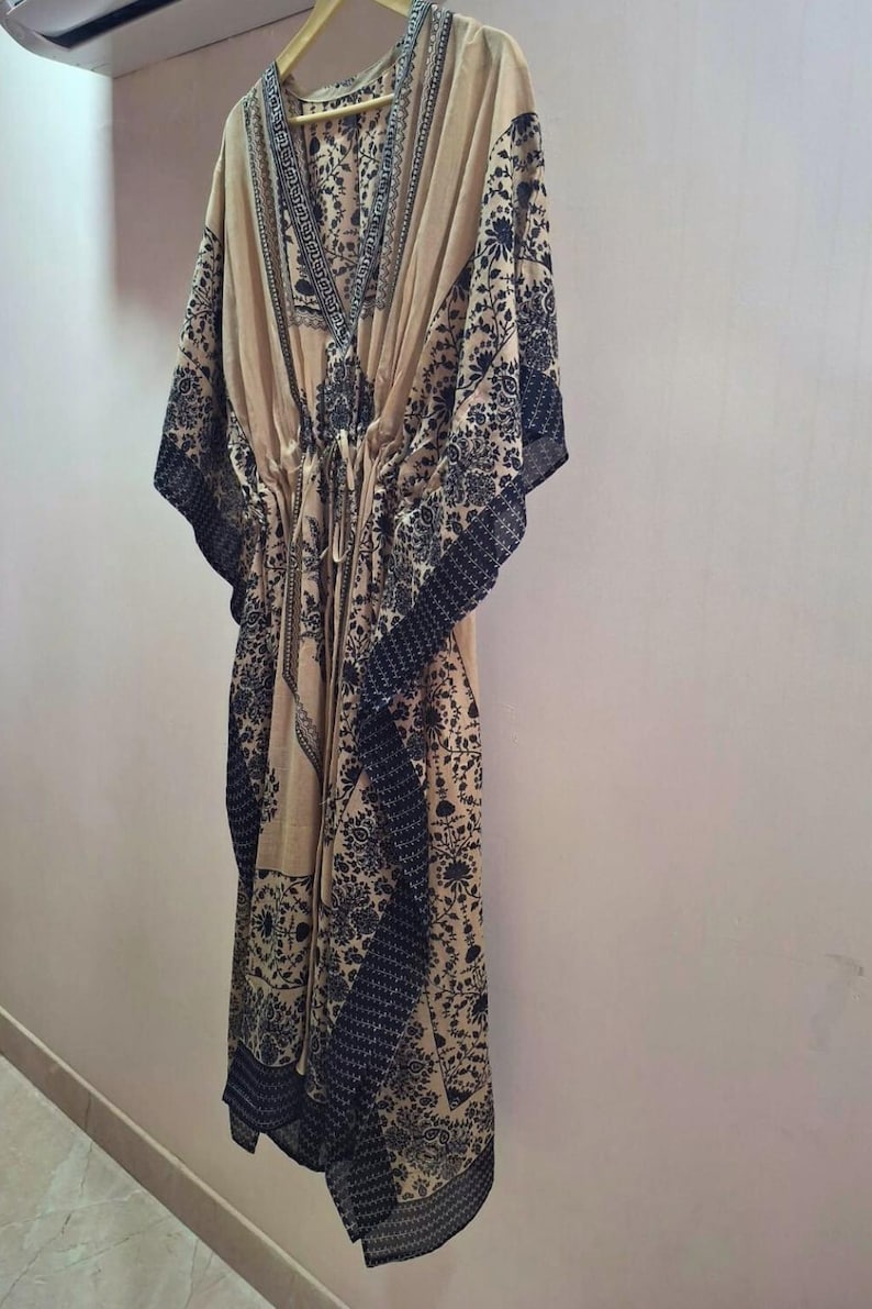 May include: A beige kaftan dress with a V-neck and a drawstring waist. The dress features a dark black floral pattern along the edges, sleeves, and neckline. It is hanging on a wooden hanger against a light-colored wall.