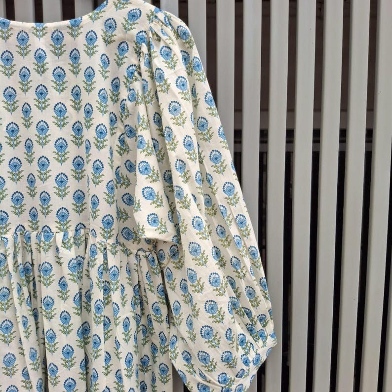 May include: A white dress with a blue floral pattern. The dress has long, puffy sleeves and a round neckline. The floral design is repeated across the fabric. The dress is made of a lightweight material.