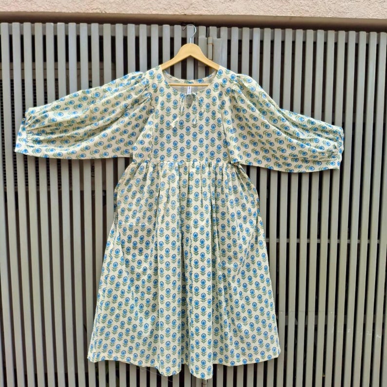 May include: A cream-colored dress with a blue floral pattern and long, voluminous sleeves. The dress is hanging on a wooden hanger against a neutral background. The dress has a gathered waist and a round neckline.