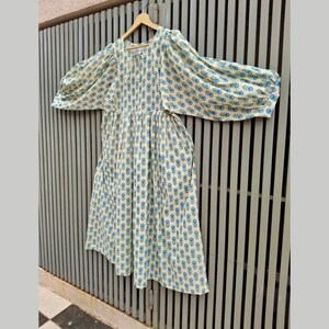 May include: A cream-colored dress with a blue floral pattern and long, puffy sleeves. The dress is hanging on a wooden hanger against a gray metal fence. The floral pattern is repeated across the entire dress.