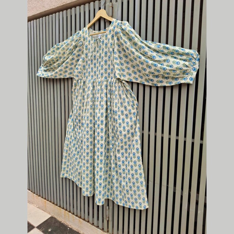 May include: A cream-colored dress with a blue floral pattern and long, puffy sleeves. The dress is hanging on a wooden hanger against a gray metal fence. The floral pattern is repeated across the entire dress.