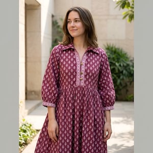 May include: A burgundy dress with a white floral pattern. The dress features a collared neckline, button-down front, and three-quarter sleeves. The fabric has a textured appearance, and the dress is gathered at the waist, creating a flowing silhouette.
