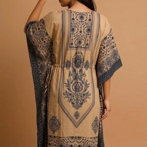 Back view of a woman wearing a beige and navy blue kaftan dress with an intricate ethnic floral print. The dress features a cinched drawstring waist, wide kimono-style sleeves, and a long midi length, set against a neutral studio background.