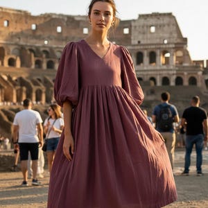 May include: A mauve-colored dress with a V-neck and puffed sleeves. The dress has a gathered skirt and falls to the mid-calf. The background features a historic building and several people. The dress is made of a soft, flowing fabric.