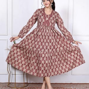 May include: A long-sleeved midi dress in a light tan color featuring a repeating dark red paisley print. The dress has a V-neckline with a decorative border, a gathered waistline, and a tiered skirt that flares out toward the hemline.