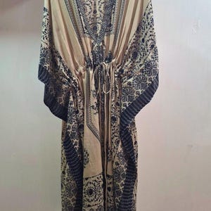 May include: A beige kaftan dress with a V-neck and a drawstring waist. The dress features a dark black floral pattern along the neckline, sleeves, and hem. The dress is hanging on a wooden hanger.