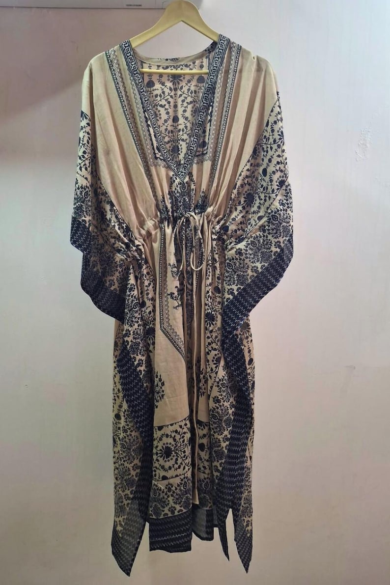 May include: A beige kaftan dress with a V-neck and a drawstring waist. The dress features a dark black floral pattern along the neckline, sleeves, and hem. The dress is hanging on a wooden hanger.