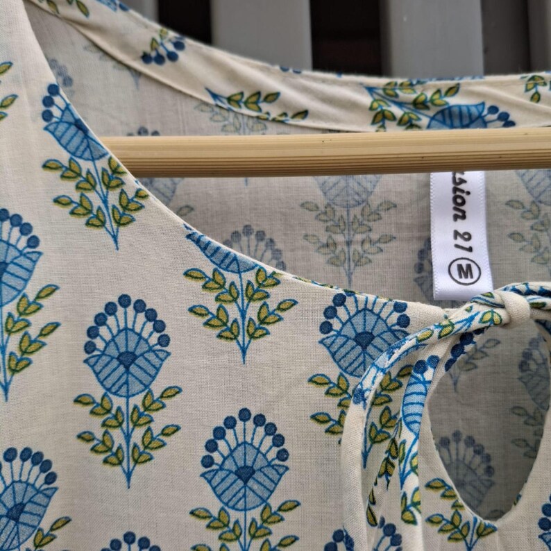 May include: Cream-colored top with a blue floral pattern and green leaves. The top has a keyhole neckline with a tie closure. The tag reads "fashion 21 M". The fabric is lightweight and appears to be cotton or a similar material.