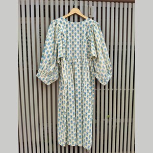May include: A long, cream-colored dress with a blue floral pattern and long, puffy sleeves. The dress is hanging on a wooden hanger against a gray and white striped background. The dress has a gathered waist and a round neckline.