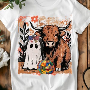 Può includere: T-shirt bianco con una mucca delle Highlands marrone e un fantasma con un fiocco con un disegno della bandiera americana. Una zucca patchwork colorata è di fronte alla mucca e al fantasma. Il design è su uno sfondo marrone e arancione.