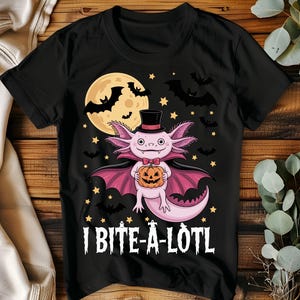 May include: Black t-shirt featuring a cartoon axolotl dressed as a vampire, holding a jack-o'-lantern. The design includes a full moon, bats, stars, and the text "I BITE-A-LOTL". A fun Halloween-themed graphic tee.