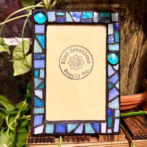 May include: A rectangular picture frame with a dark border decorated with a mosaic of blue, light blue, and turquoise glass pieces. The center insert features a circular logo with the text Kind Creations Supply Co. and a floral icon.