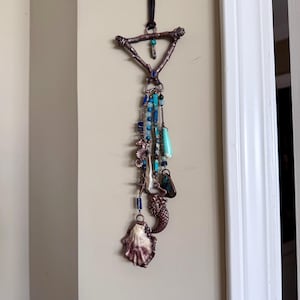 May include: A handcrafted wall hanging featuring a triangular, twig-like frame. Turquoise, blue, and clear beads dangle below, along with a seashell, mermaid charm, and other decorative elements. The piece is suspended by a dark brown cord.