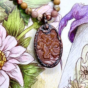 May include: A beaded necklace featuring a pendant with a carved design. The pendant is a reddish-brown color with a darker border. The beads are a mix of pink, brown, and tan tones. The background is a floral illustration.