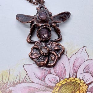 May include: A copper-coloured pendant necklace with a bee design. The bee has outstretched wings and holds a flower. The pendant incorporates three oval-shaped, purple gemstones. The necklace chain is also copper-coloured.