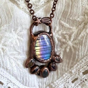 Op de afbeelding: Een koperen ketting met een regenboogkleurige, iriserende edelsteenhanger en kleinere edelstenen rondom de omtrek. De hanger heeft de vorm van een traan en heeft een stervormige hanger aan de bovenkant.