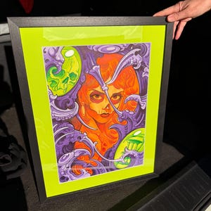 May include: Framed art print featuring a stylized portrait of a woman with orange hair and a skull. The artwork is set against a neon green background and framed in black. The image has purple and green decorative elements.