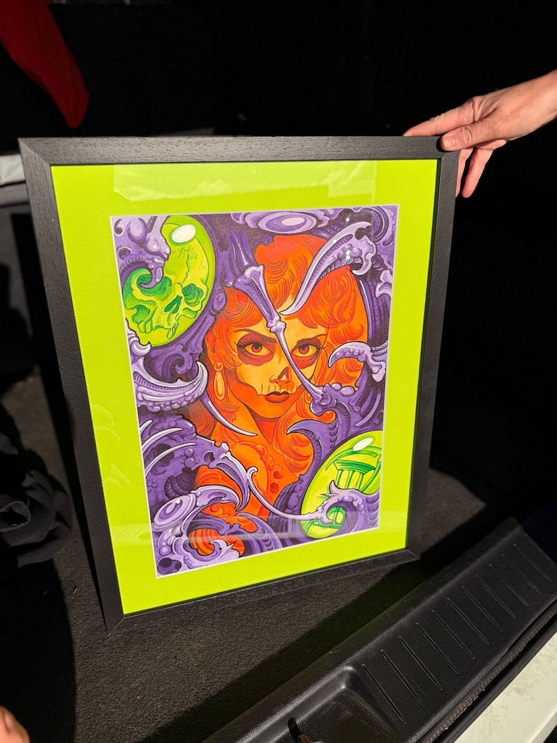 May include: Framed art print featuring a stylized portrait of a woman with orange hair and a skull. The artwork is set against a neon green background and framed in black. The image has purple and green decorative elements.