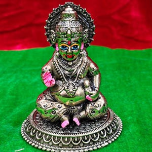 May include: A detailed bronze-colored statue of a seated deity with intricate jewelry and a halo. The figure has blue eyes and pink accents on the hands and feet. It sits on a decorative, circular base against a red and green background.